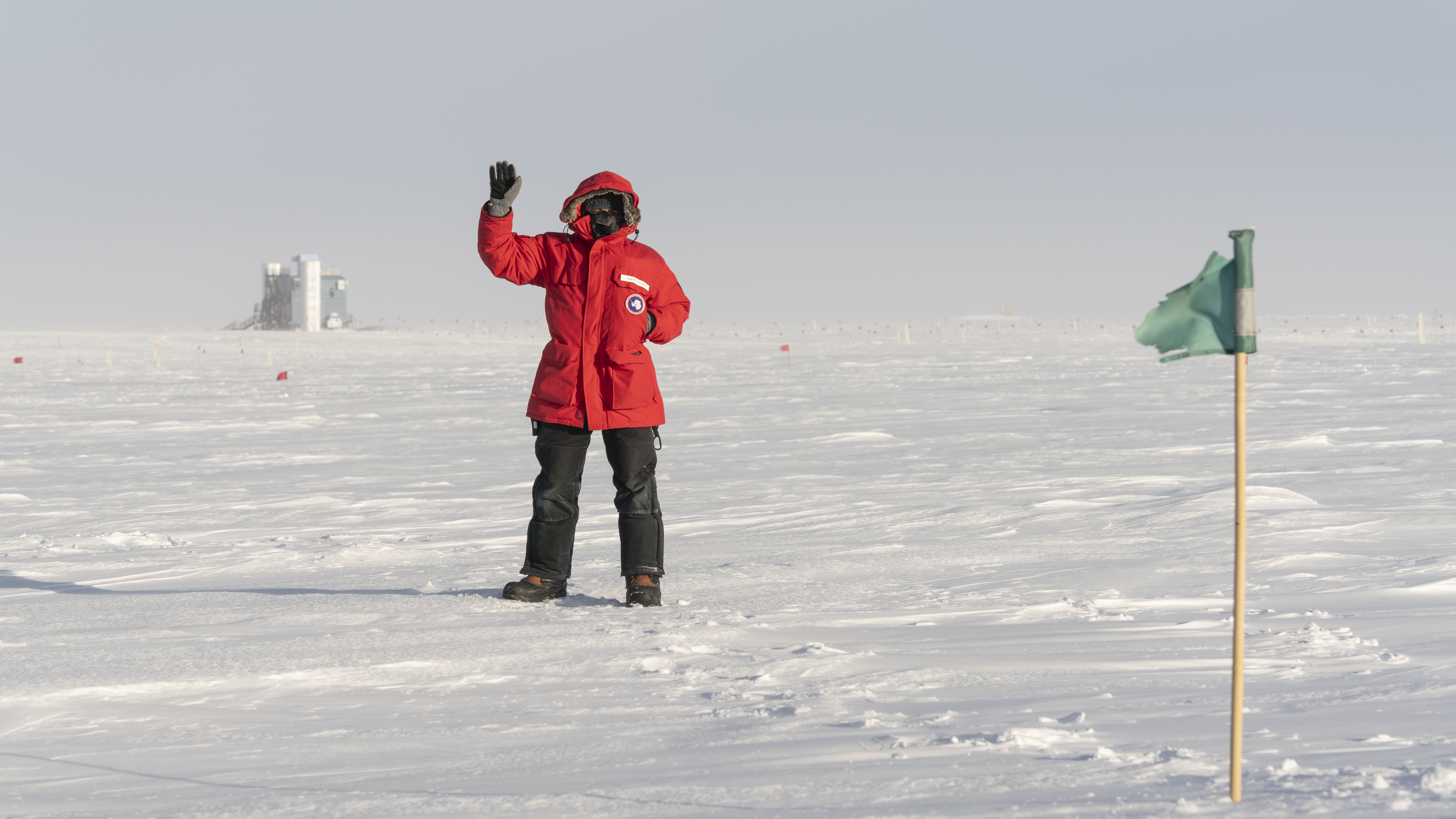 Person in red parka waving, with the IceCube Lab in the distance.