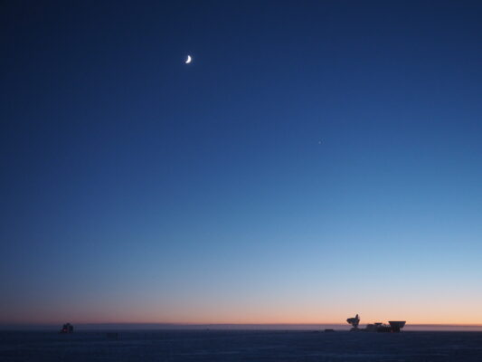 Twilight at the South Pole, with crescent moon high in sky and orange hues along horizon.