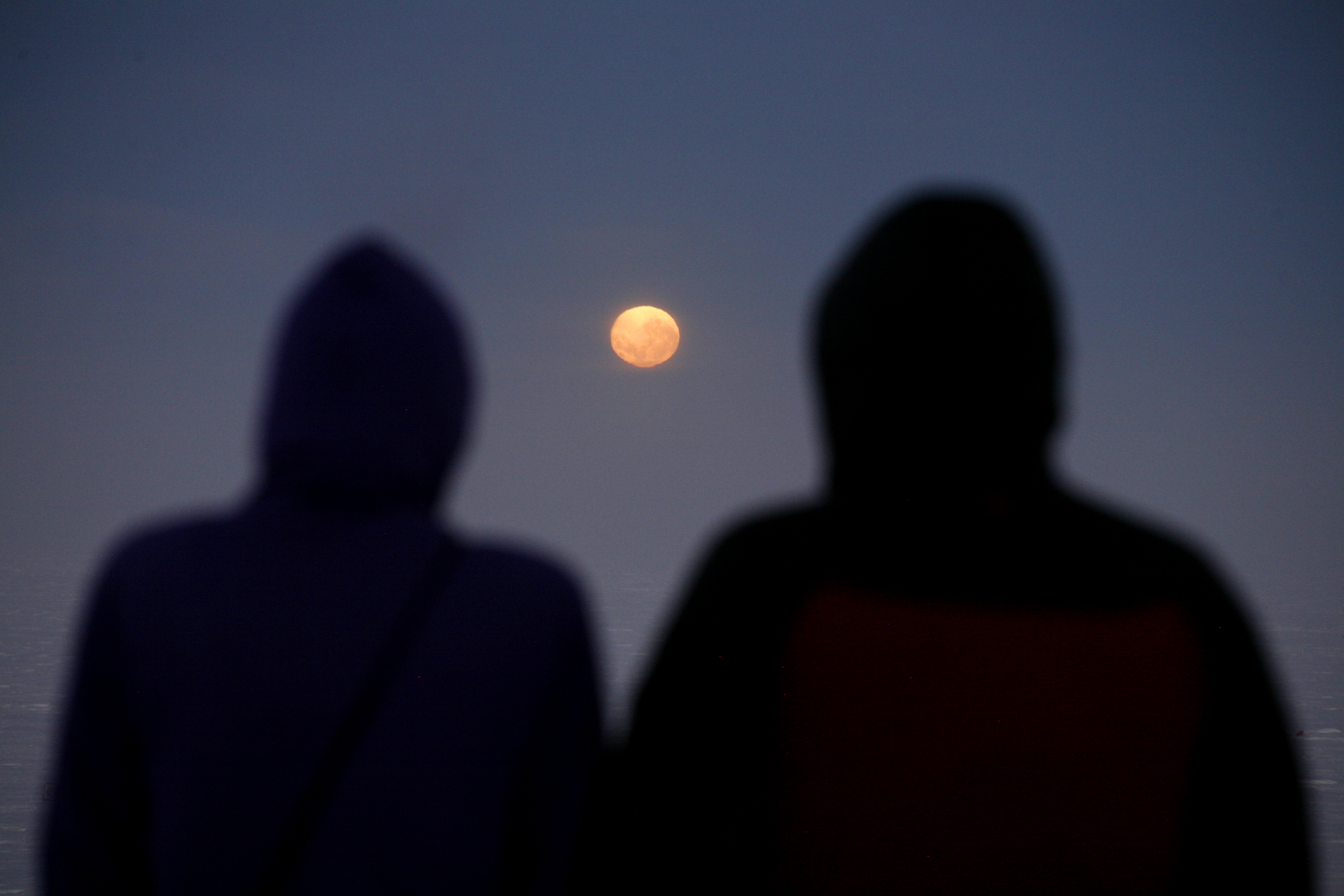 Blurry images of shadowed behinds of two hooded individuals facing a full moon, shown between them.