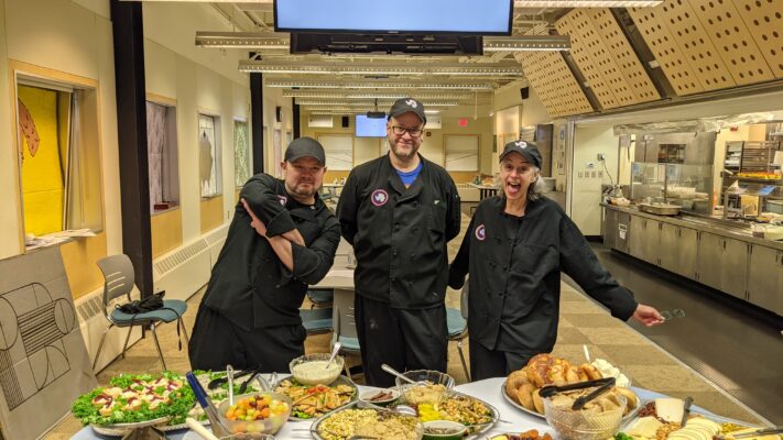 Three galley staff posing behind table set up with brunch food.