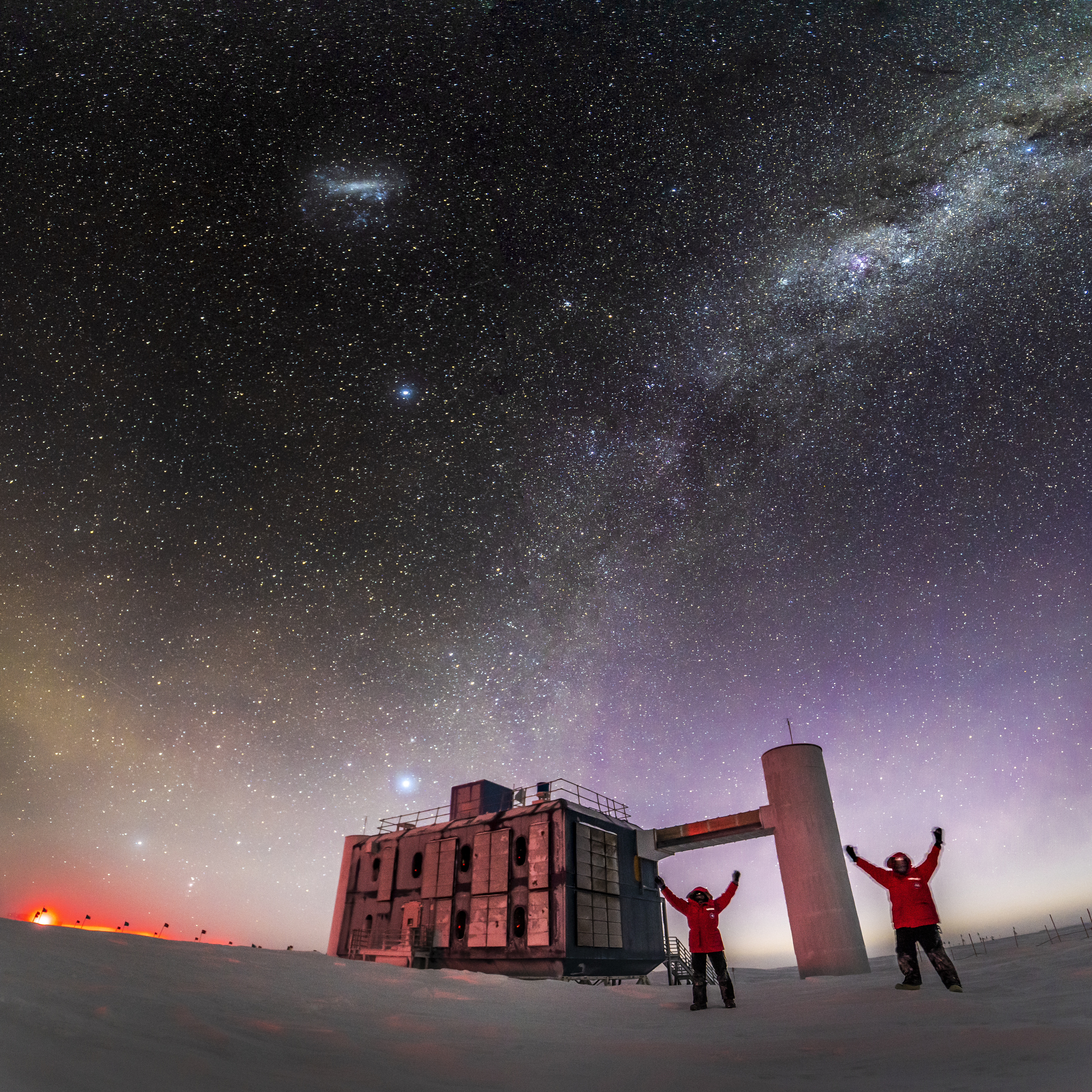 Winterovers Martin and Josh taking selfi outside the IceCube Lab, with arms up, stars in sky and sunlight along horizon.