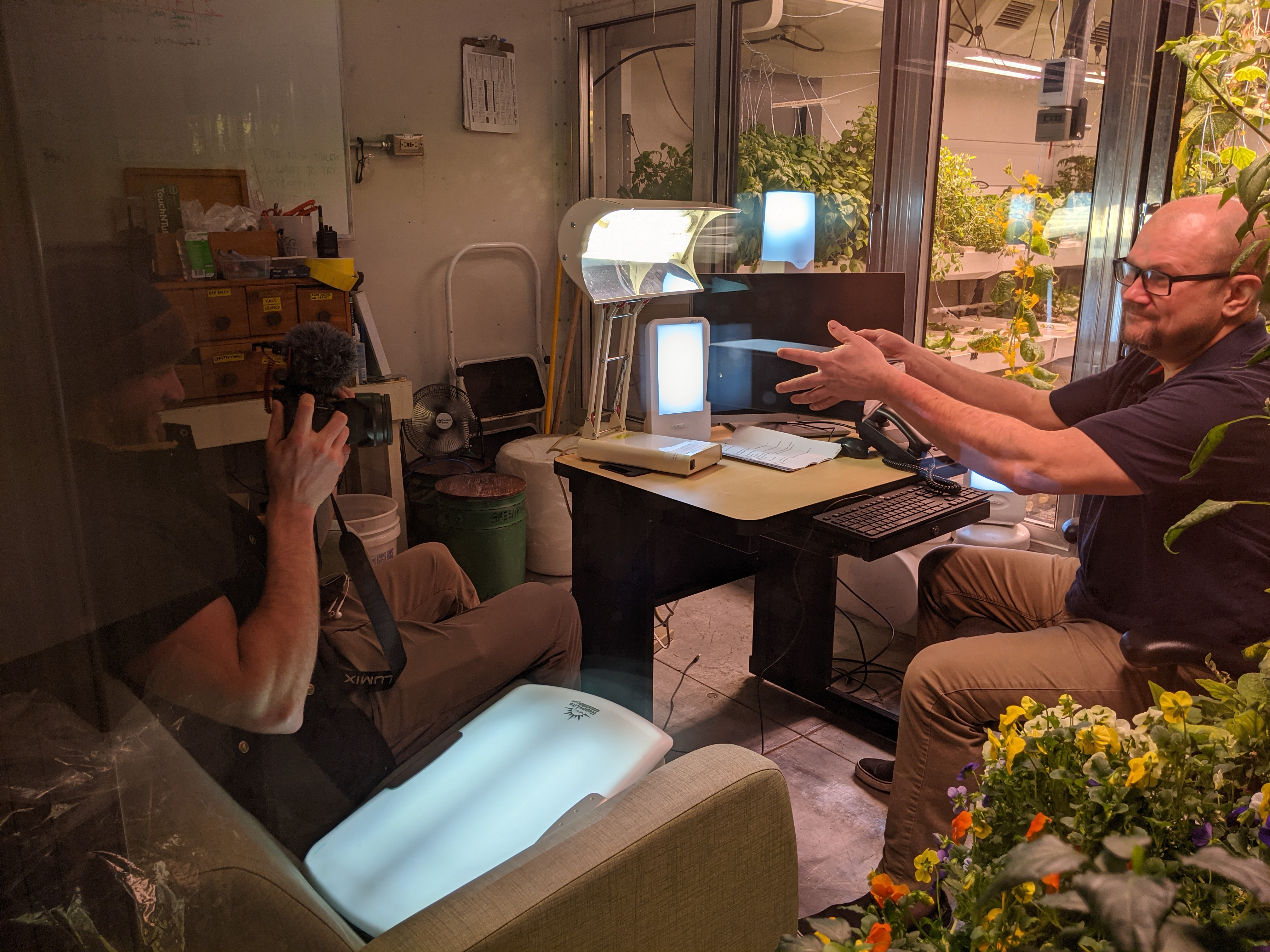 Two people seated in an office space, one filming the other.