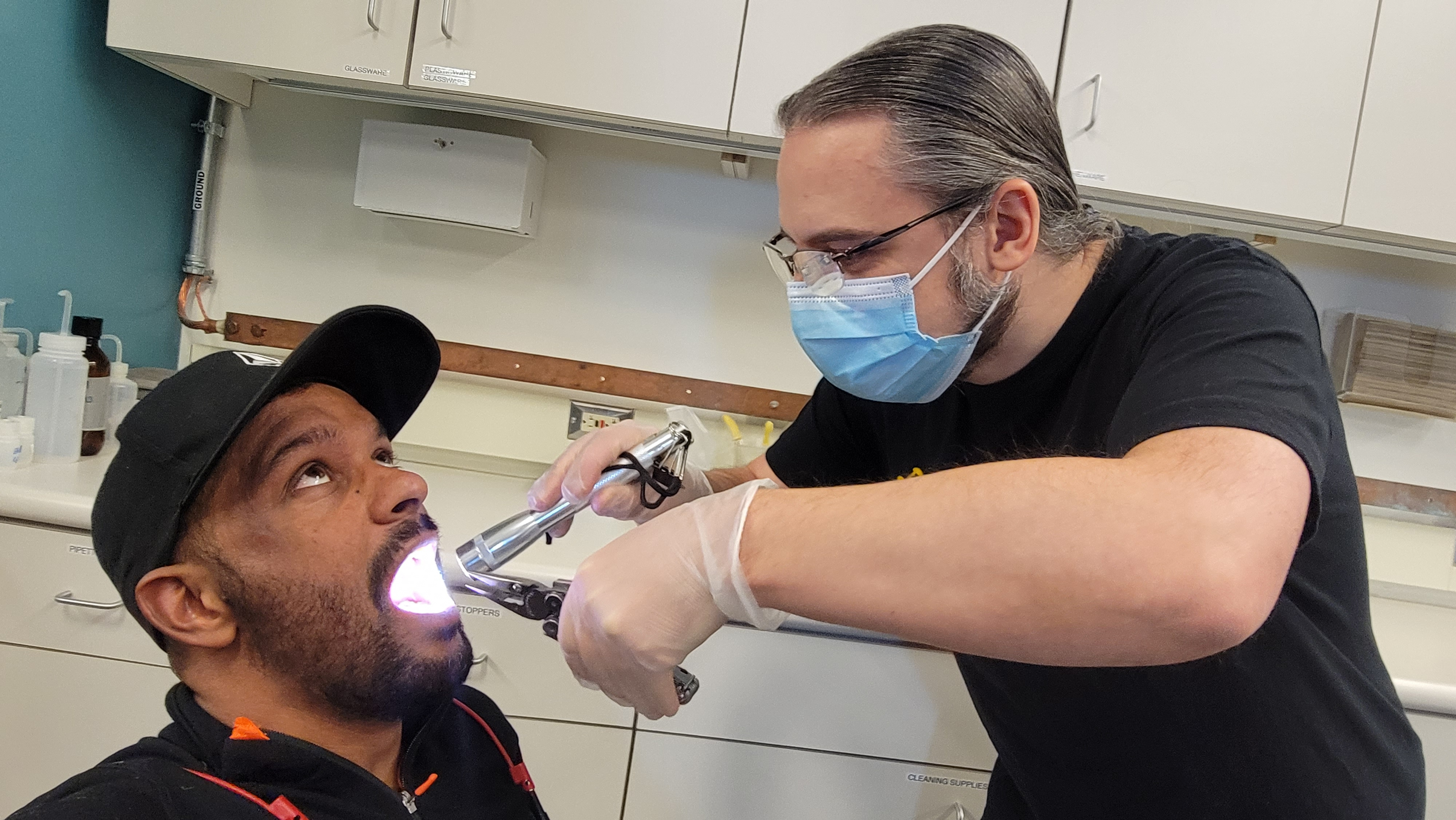 Close-up of two people posing as dentist and patient, one wearing surgical mask and holding flashlight and another tool while peering into the other’s open mouth.