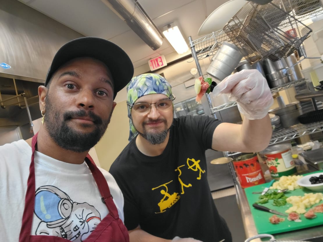 Two people in kitchen, facing camera with cutting board off to side and one person holding up skewer.