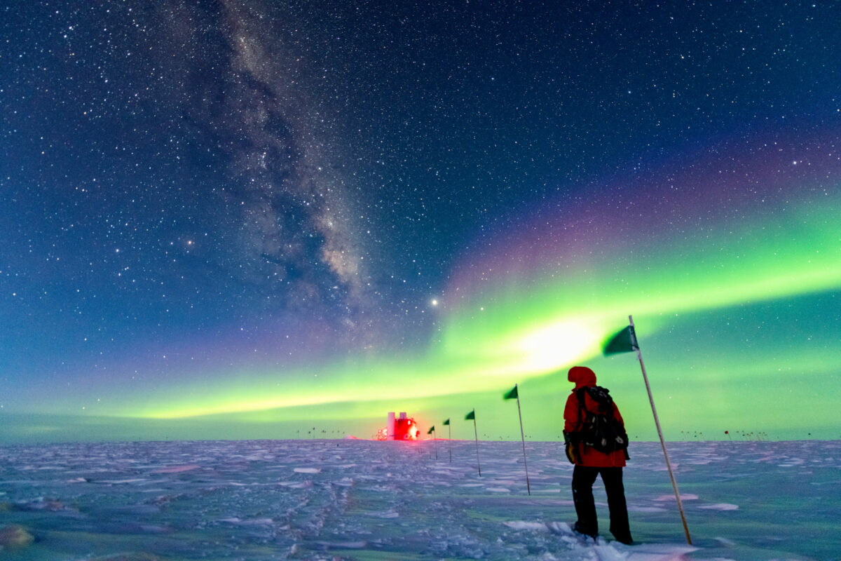 A winterover walking along a flag line in the direction of the IceCube laboratory in the distance in the South Pole. The night sky above is filled with the aurora australis, the galactic plane, and the Milky Way.