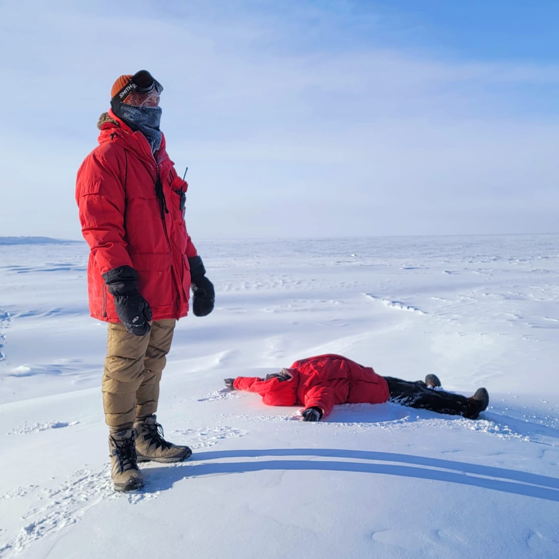 Winterovers watching air drop, one standing upright the other lying face up on the ice.