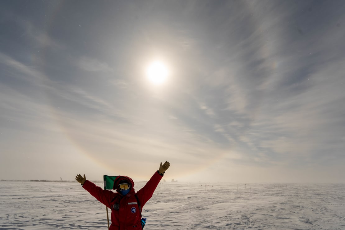 Person in red parka with arms up in air, below a sun halo.