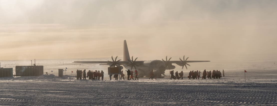 Group of people boarding a large plane in the distance on the ice at the South Pole under somewhat hazy sky.