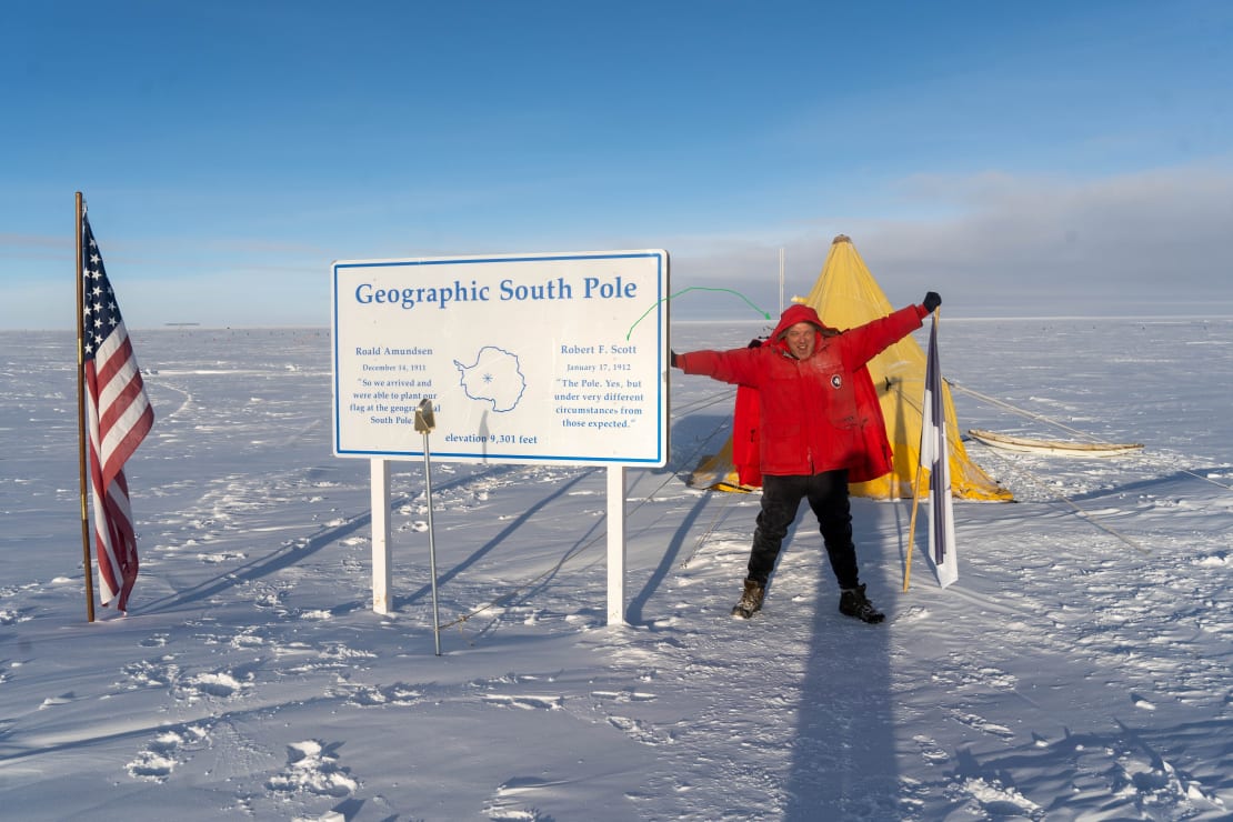 Winterover in red parka with arms outstretched, posing in front of a pitched Scott tent at geographic South Pole.