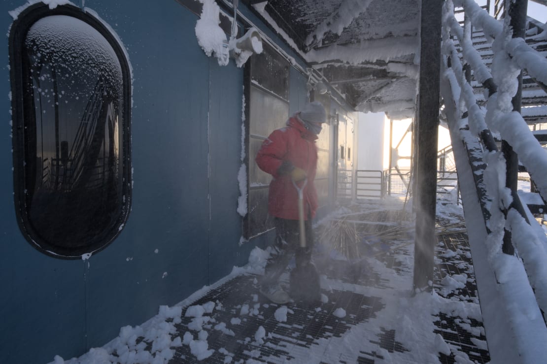 Winterover in red parka standing on deck of the IceCube Lab after clearing off accumulated snow.