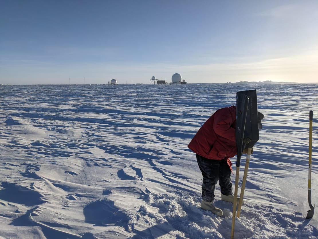 A winterover in red parka installing a flag into the snow at the South Pole, satellite communications structures in background along horizon.