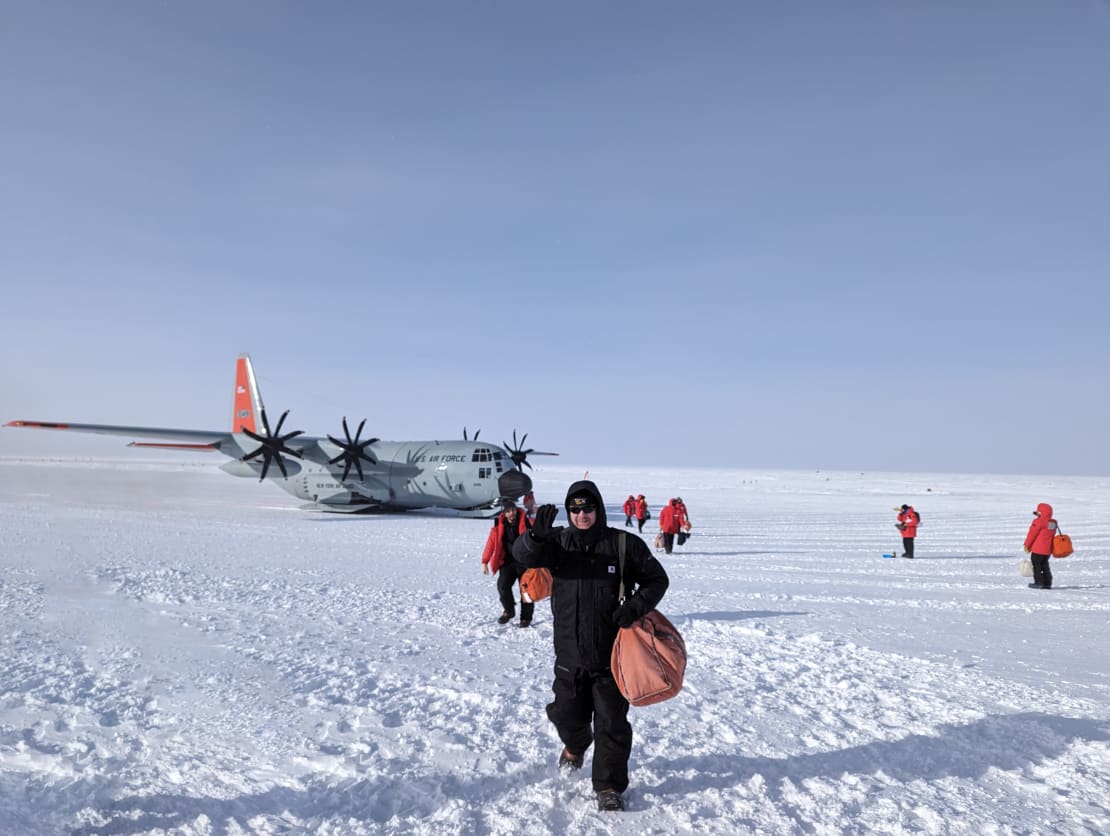 A large plane parked on the ice in the background, with some people in parkas walking toward and away from the plane.