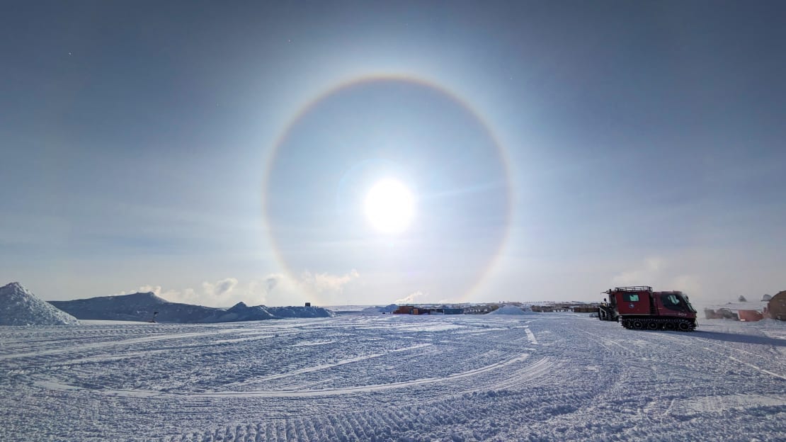 A bright sun with a halo at the South Pole, with some snow mounds and snow vehicles off to the side.