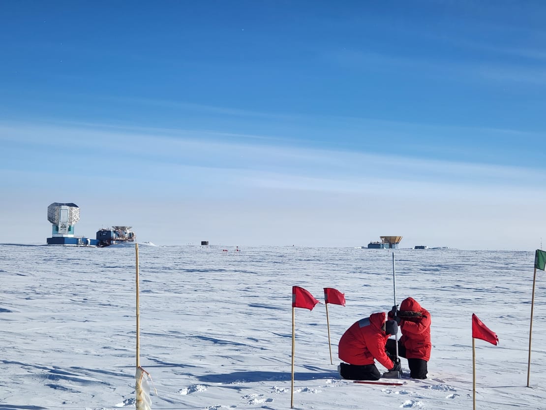 Two winterovers in red parkas crouched down on ice under blue skies while taking snow depth measurements.