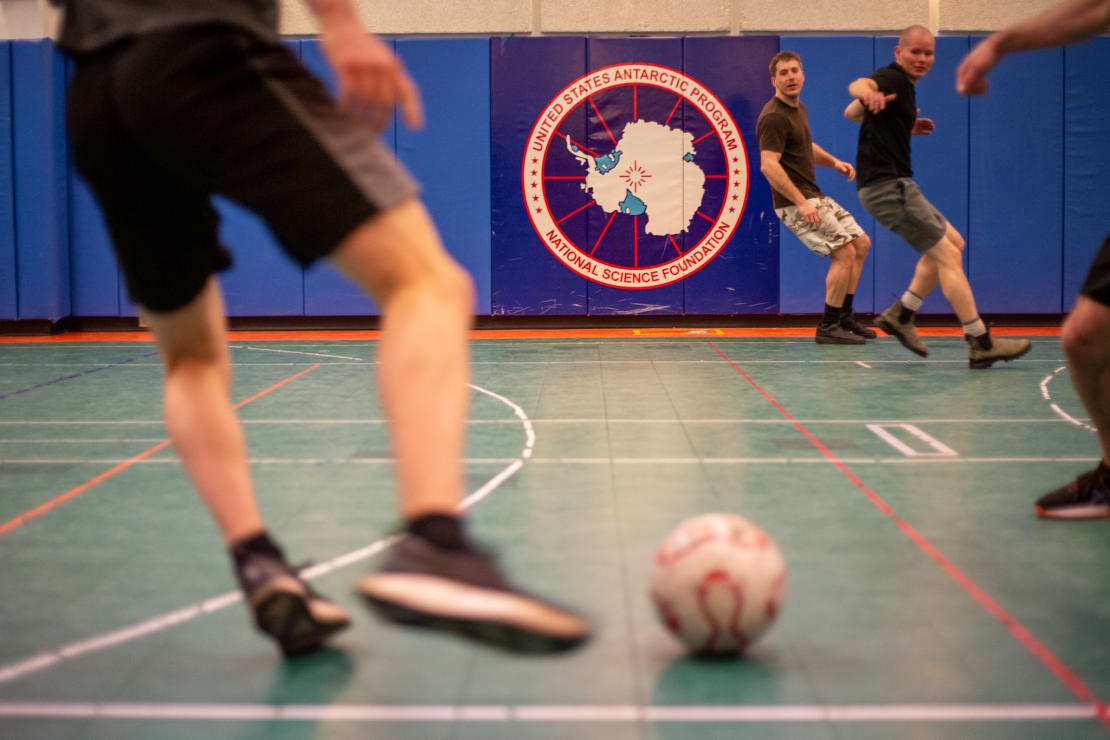 Action shot in gym, with out-of-focus close-up from behind of soccer ball about to be kicked, focus on players down field about to react.