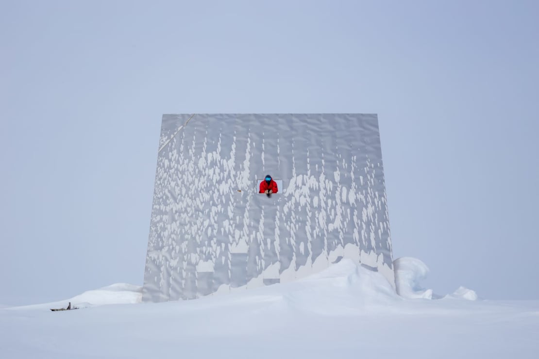 A huge reflector paneal at the South Pole, snowswept and with gray skies behind, and a winterover poking thru a small window about two-thirds the way up from the ground.