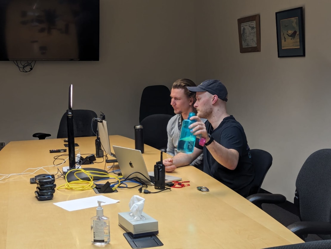 Two people participating in a video call, viewed from the side, seated in front of monitors at conference room table.