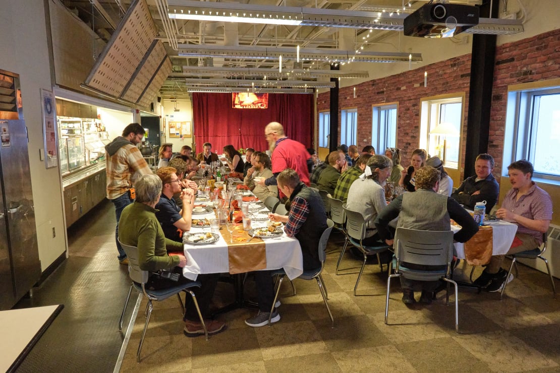 Long view of two tables filled with people sitting down to sunset dinner in the South Pole station galley.