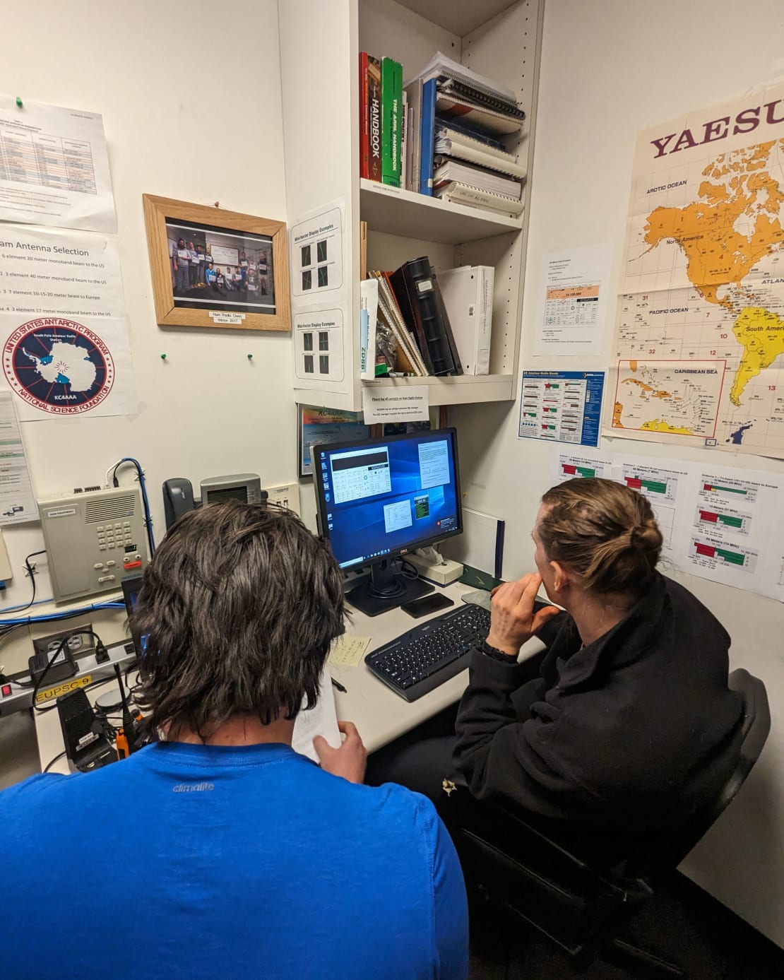 Two people seen from behind while seated at a desk with radio equipment and computer monitors.