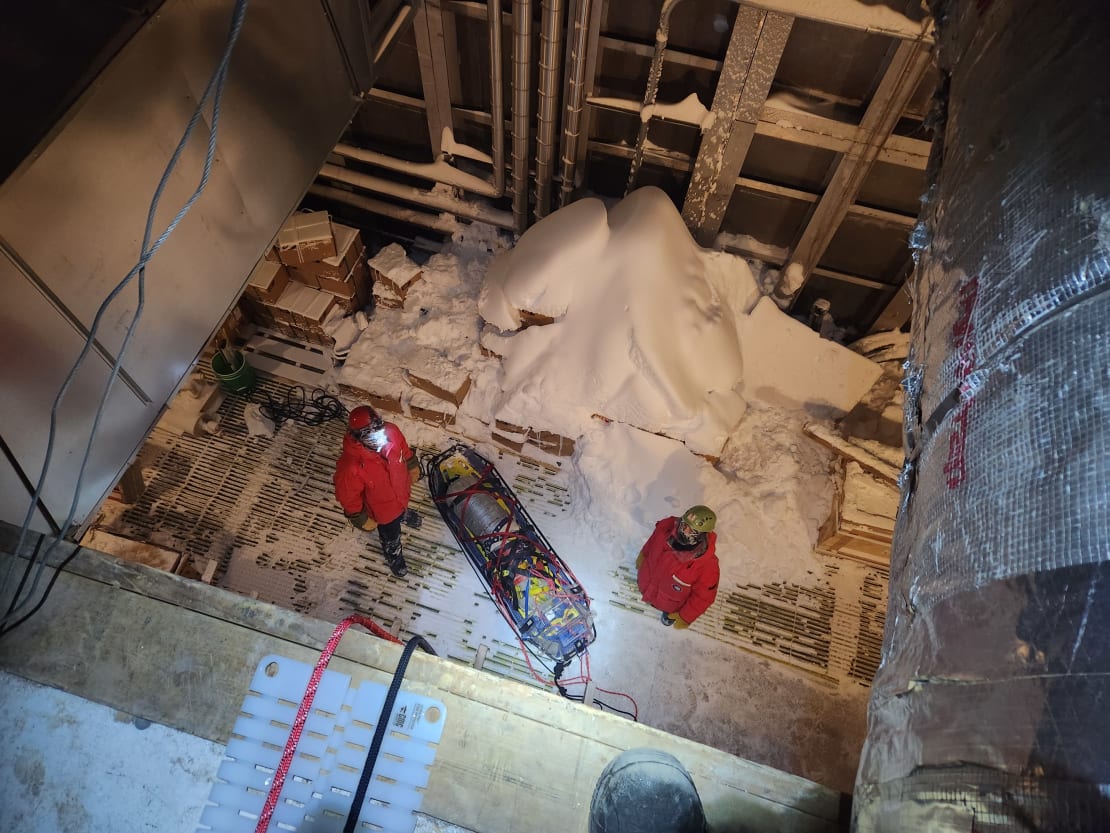 View from above at two people in red parkas, looking up, about to hoist up a dummy rescue rig.