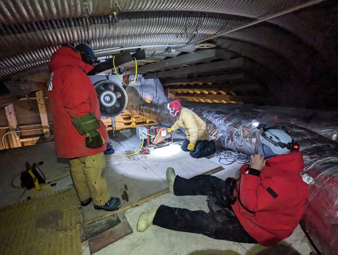 Several people in red parkas or cold weather gear using headlamps in darkened fuel arch under the South Pole station.