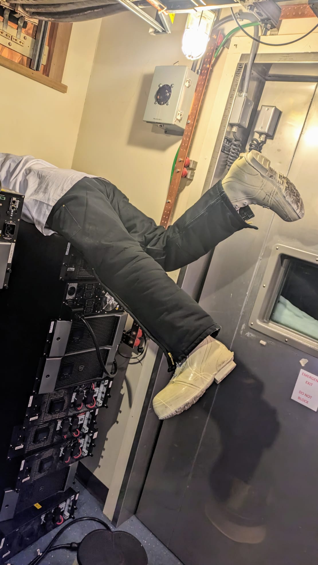 Backside of person, with legs kicking, as they’ve hoisted themselves up onto stack of computers to read a serial number.