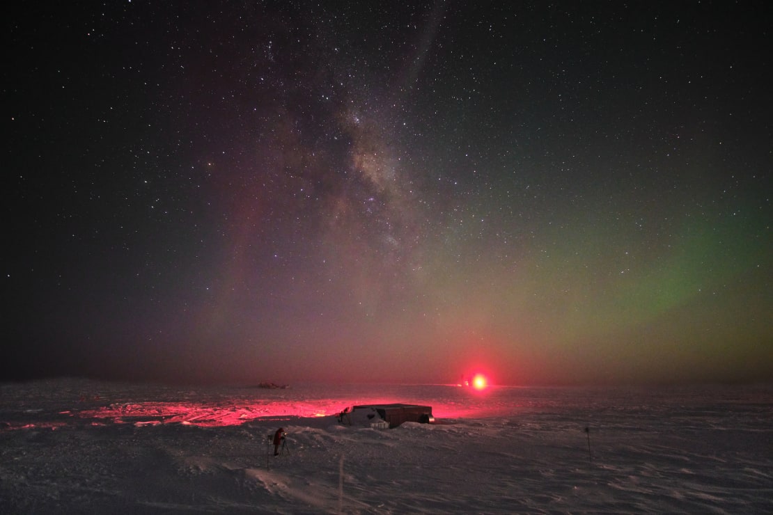 The Milky Way along with some faint auroras in the South Pole sky, with a bright red light on the horizon.