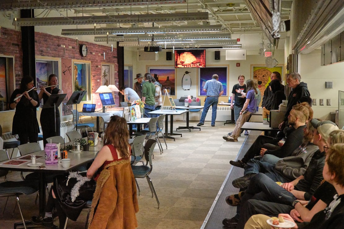 Room set up with tables displaying art, two people playing flutes, other people milling about or seated along perimeter.