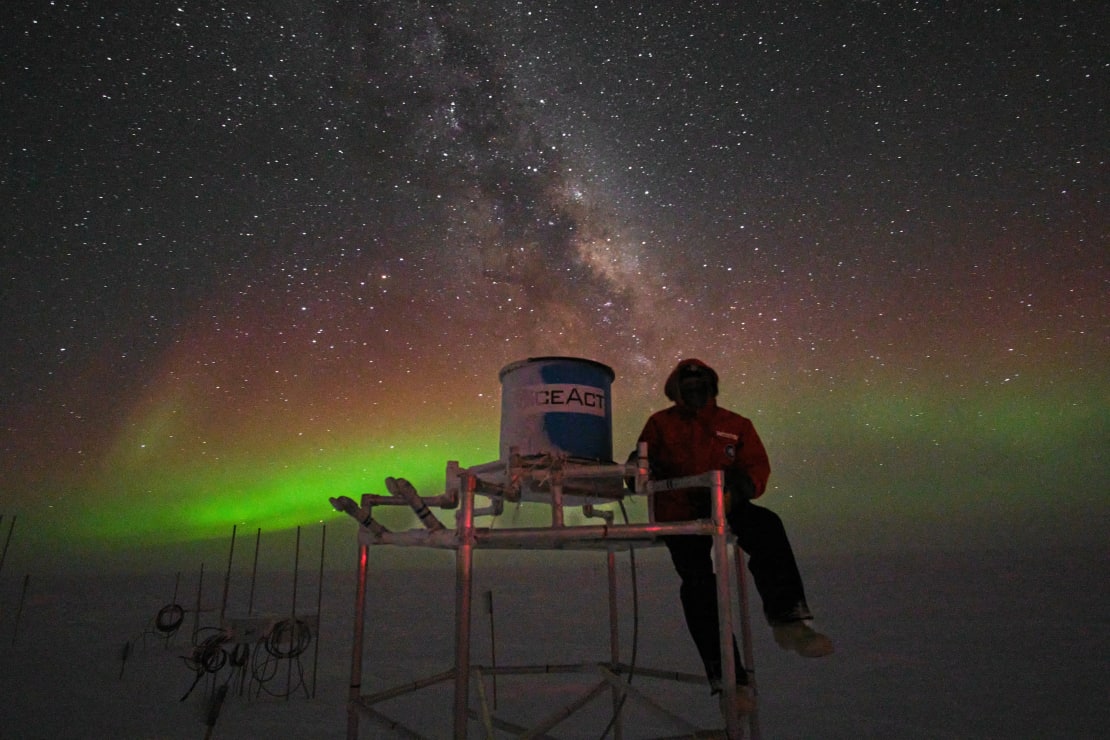 Winterover posing next to IceAct telescope under winter sky filled with stars and auroras.