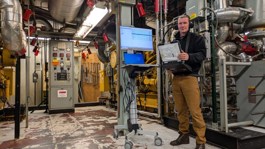 Inside view of South Pole power plant, person standing off center wearing ear mugs and holding laptop.