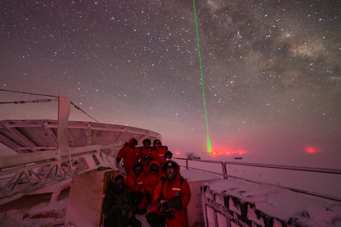 Group in red parkas gathered for photo at the South Pole Telescope in winter, starry skies as backdrop.