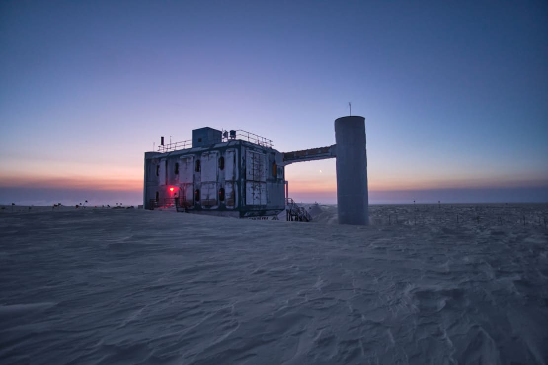 Angled view of frosty IceCube Lab in shadow during early sunrise, small faint moon just visible above horizon.