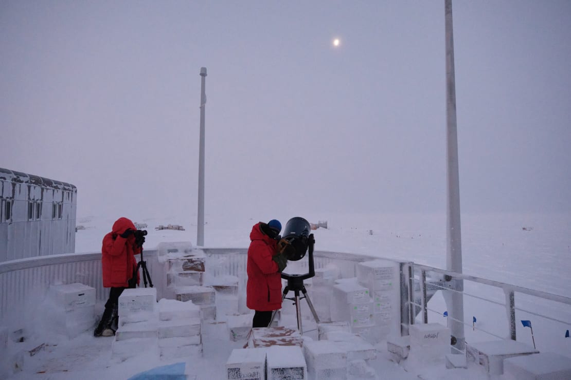 Two people in red parkas on South Pole observation desk looking through telescopes at the moon.