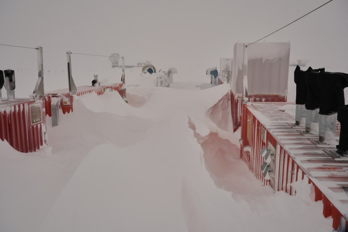 Tall snow drift against red container structures at the South Pole.