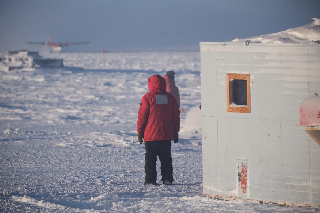 A twin otter, blurry in the far distance, with the backs of two onlooking winterovers in red parkas in the foreground.