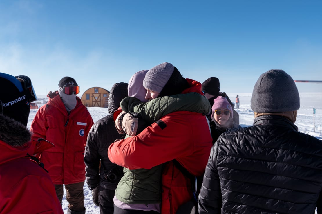 Group of winterovers out on the ice, focus on side view of two winterovers embracing before one departs the Pole.