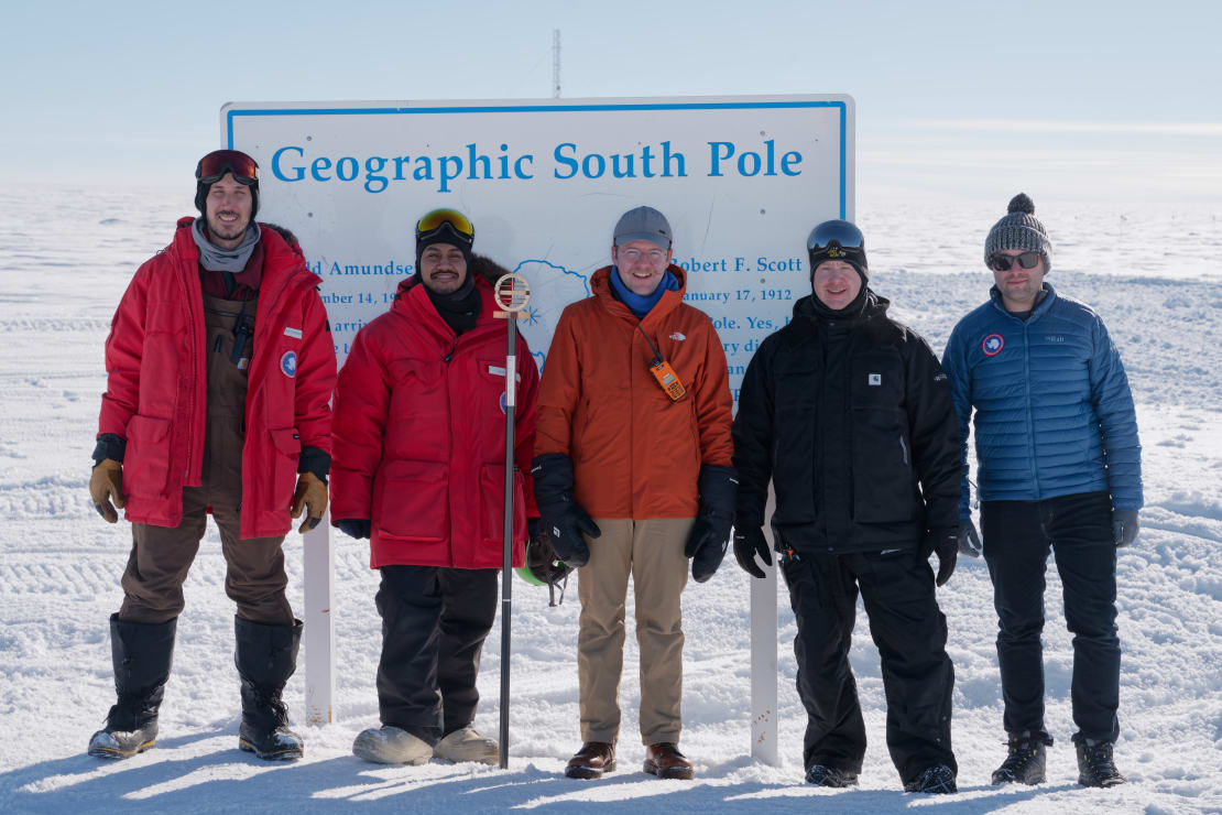 Five people posing in front of the geographic South Pole sign.