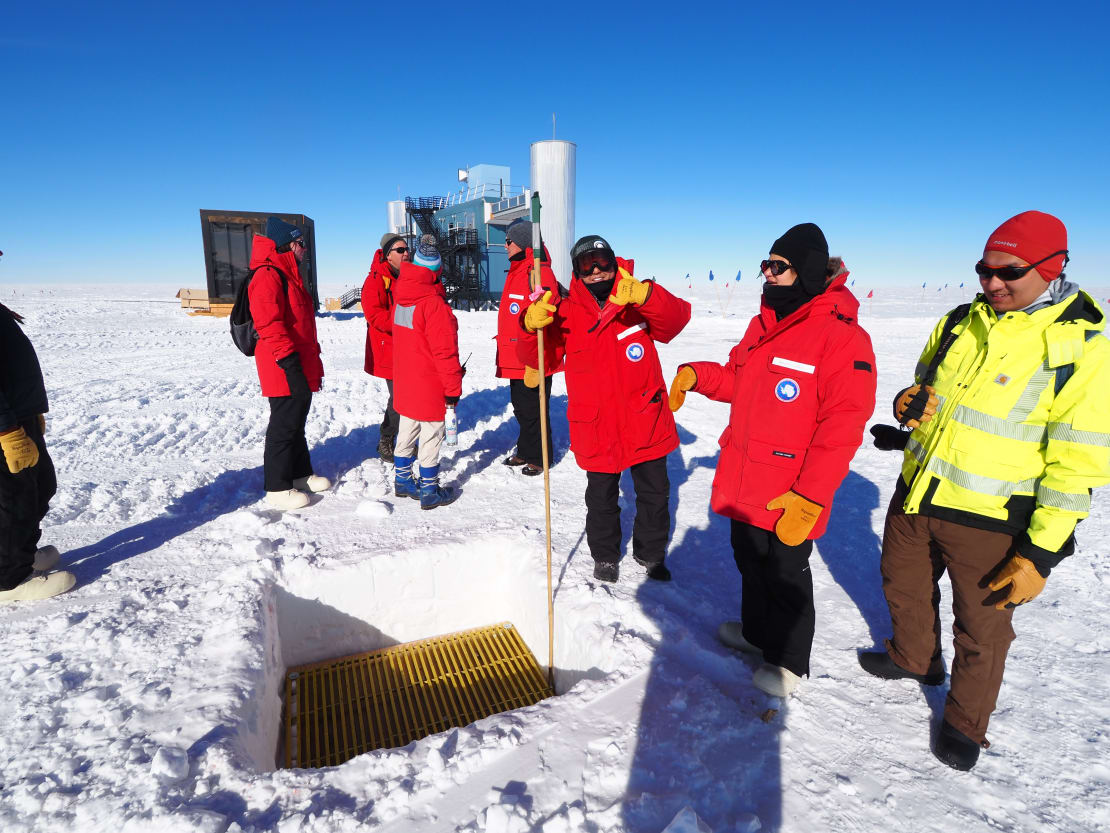 A group of people in parkas, outside, near a dugout spot marking the next drill hole site, IceCube Lab in the background.