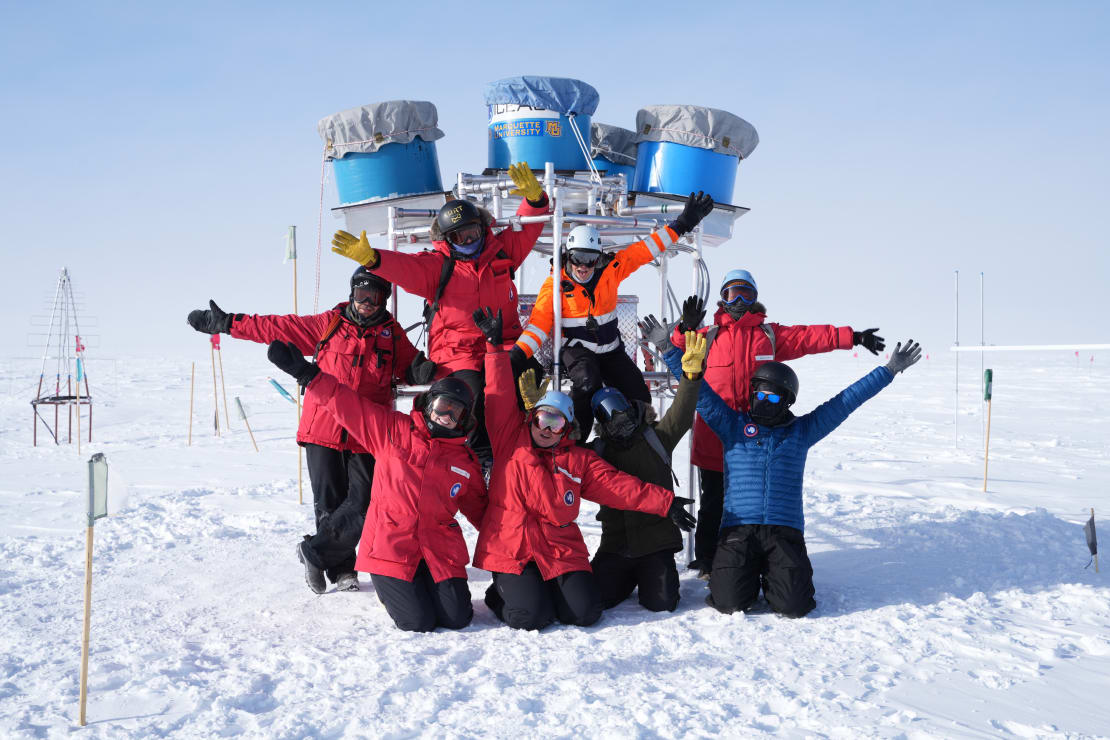 Group of people posing outside at the South Pole, arms up in the air, positioned in front of scaffolding with large blue cylindrical structures on top.