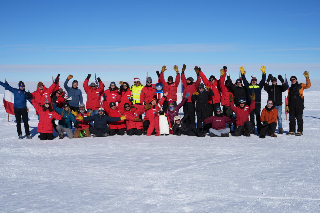 Large group photo outside at the South Pole on bright day, many with arms raised high.