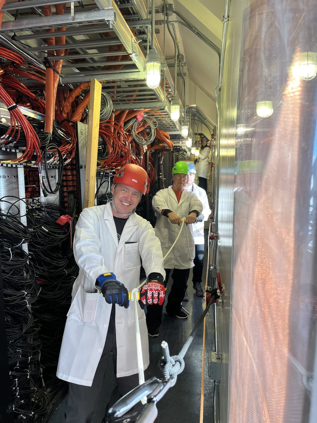 Group of people in lab coats and hard hats facing forward, all holding onto a cable, ready to pull.