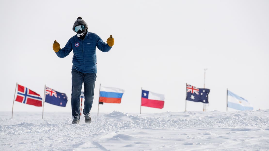 South Pole marathon participant giving thumbs up with both hands out on the ice, some of the flags at the ceremonial Pole low in the backhground.