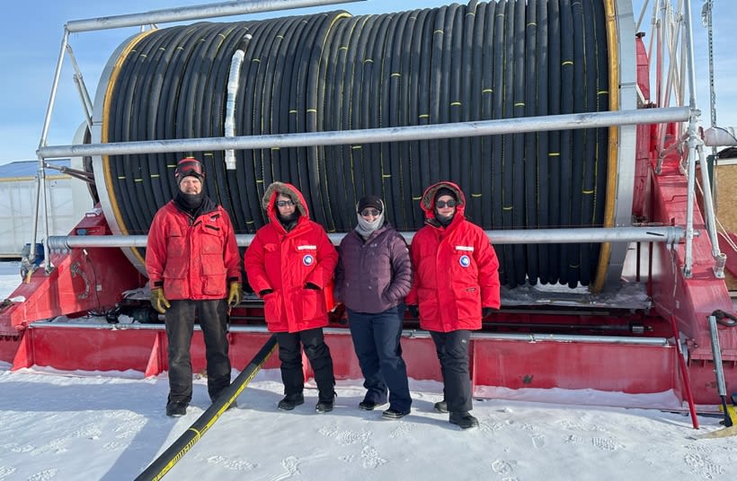 A group of people in red coats standing in front of a big reel loaded with a hose