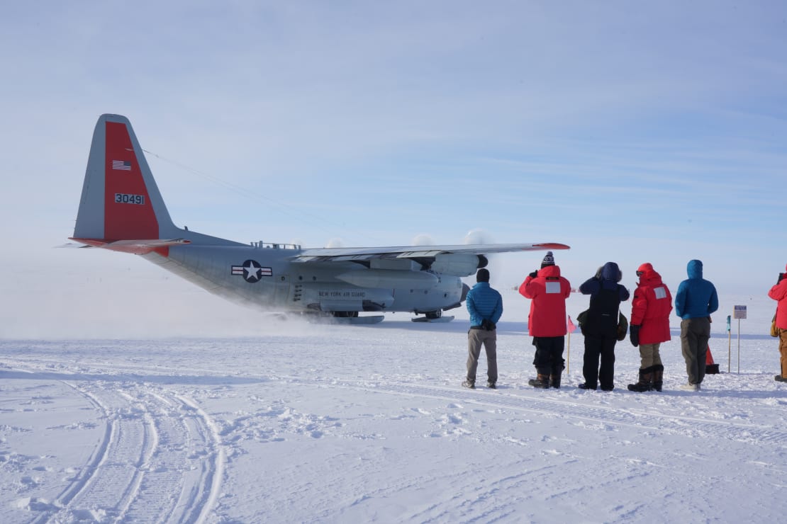 Small group of people lined up, seen from behind, out on the ice looking on as LC-130 plane leaves the South Pole