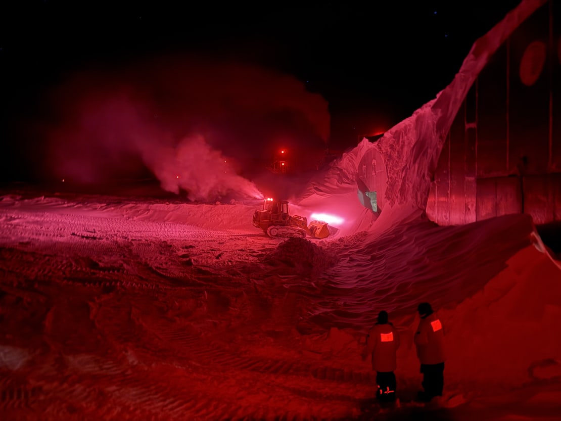 Heavy equipment removing snow from South Pole storage arches in darkness under red lights.
