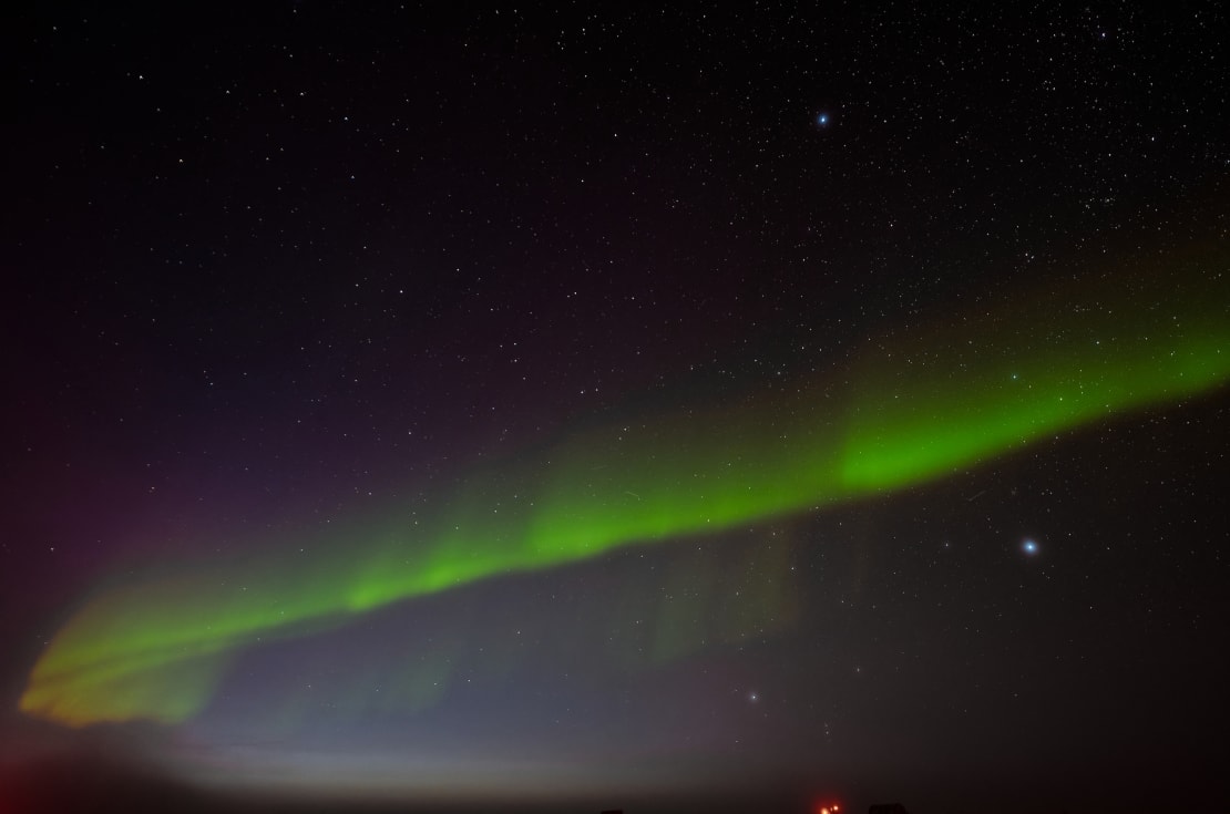 Bright green streak of aurora spreading as diagonal line across full star-studded sky.