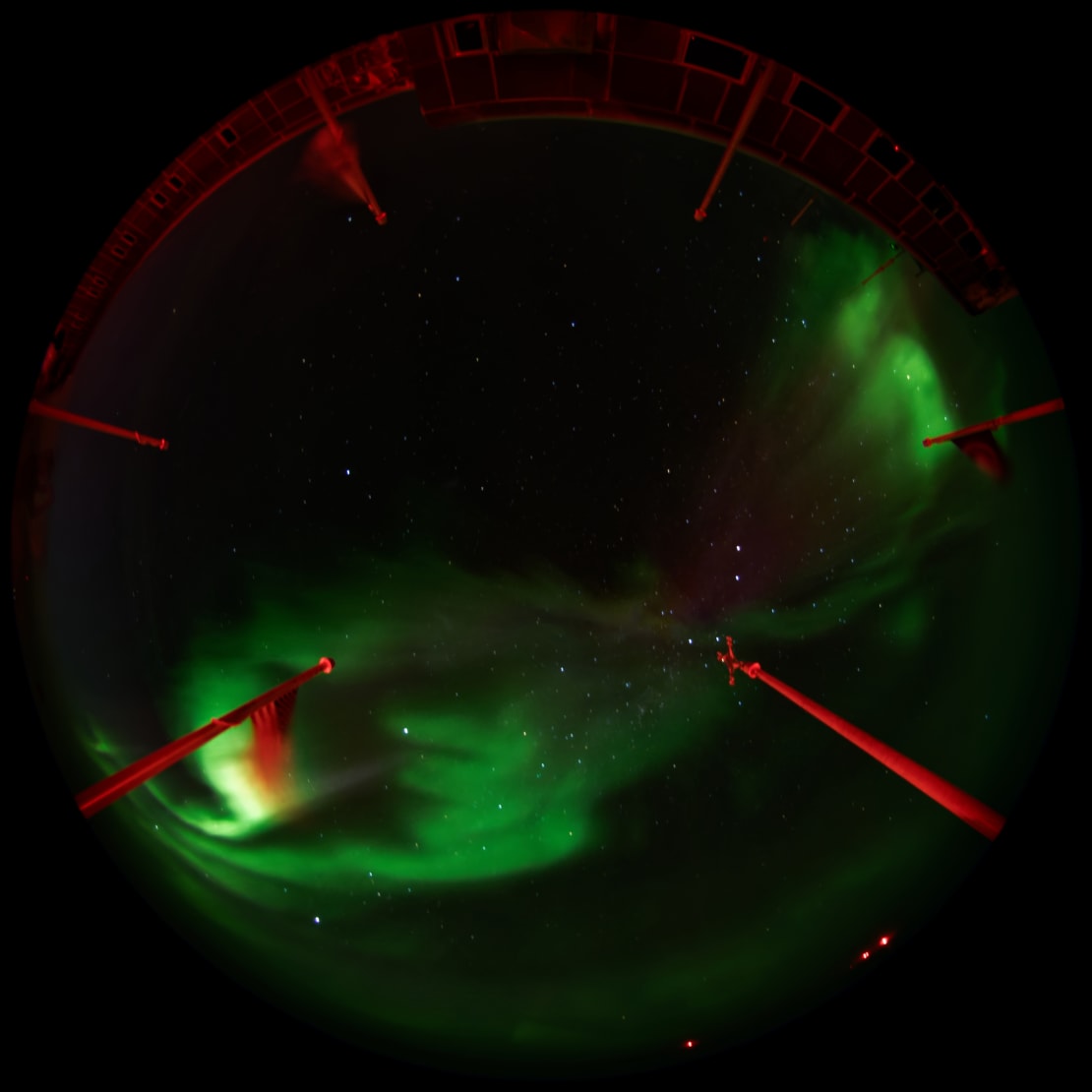 A fisheye view of green swirling auroras from the South Pole station’s observation deck, with flag poles lit in red.