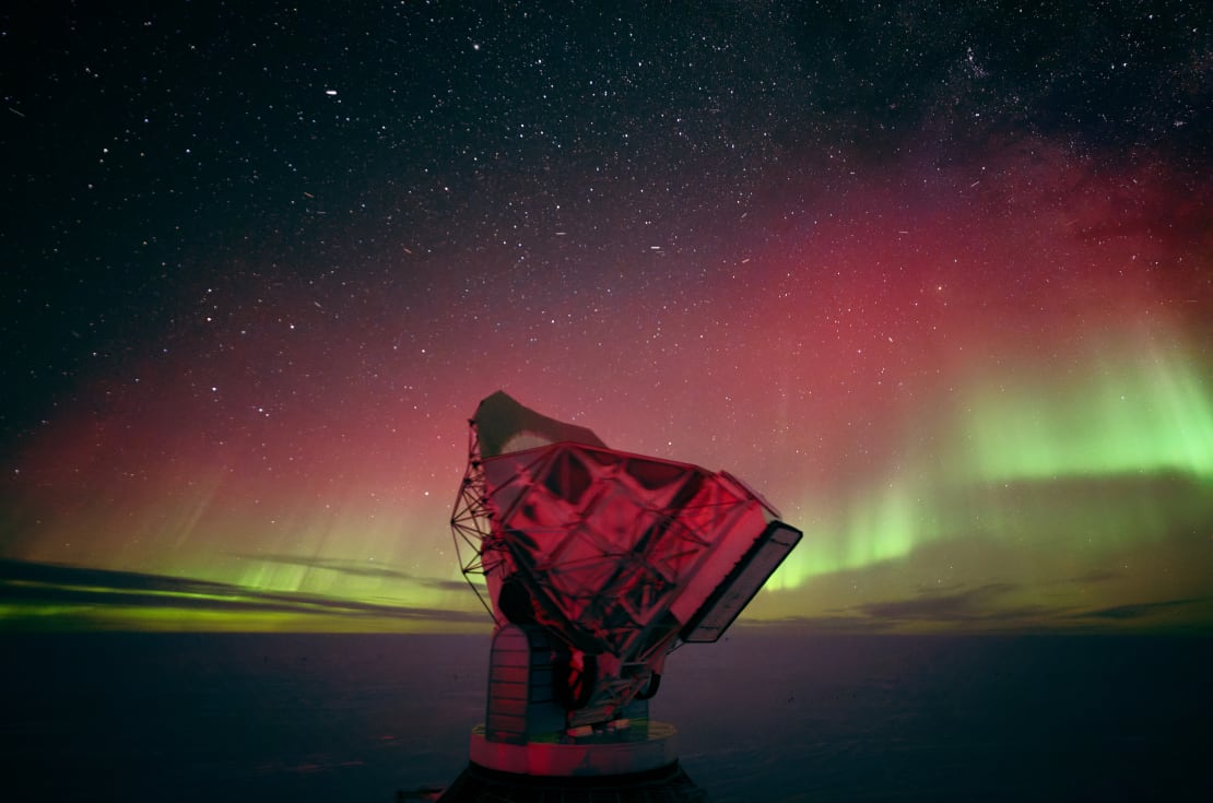 Beautiful green and red auroras stretching upward from low on the horizon at the South Pole, with the South Pole Telescope barely lit in red in the foreground.