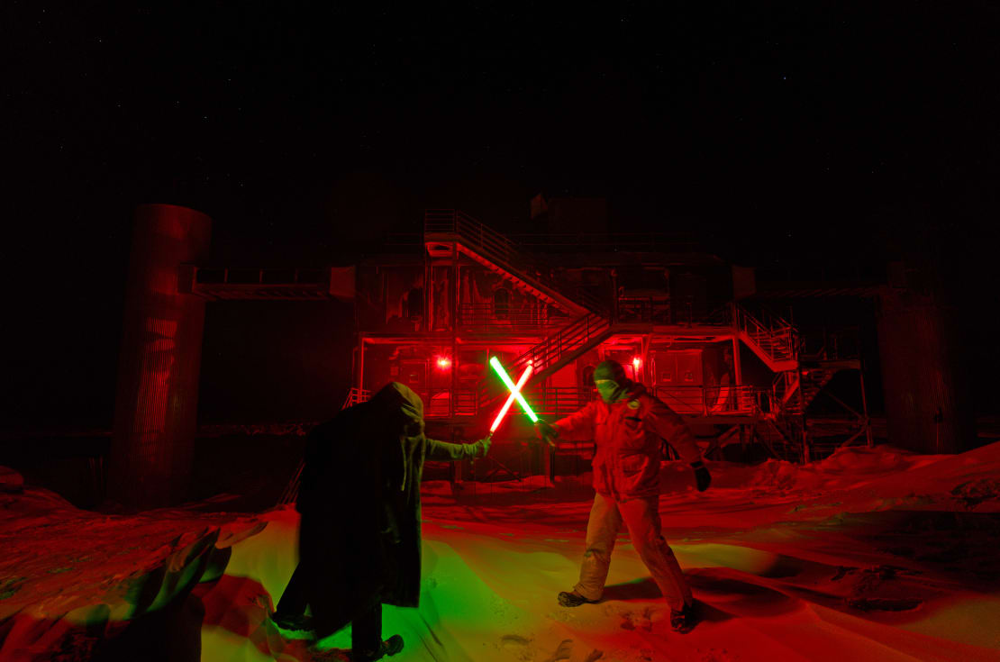 Winterovers facing off with lightsabers, which form a bright X, in front of the IceCube Lab.