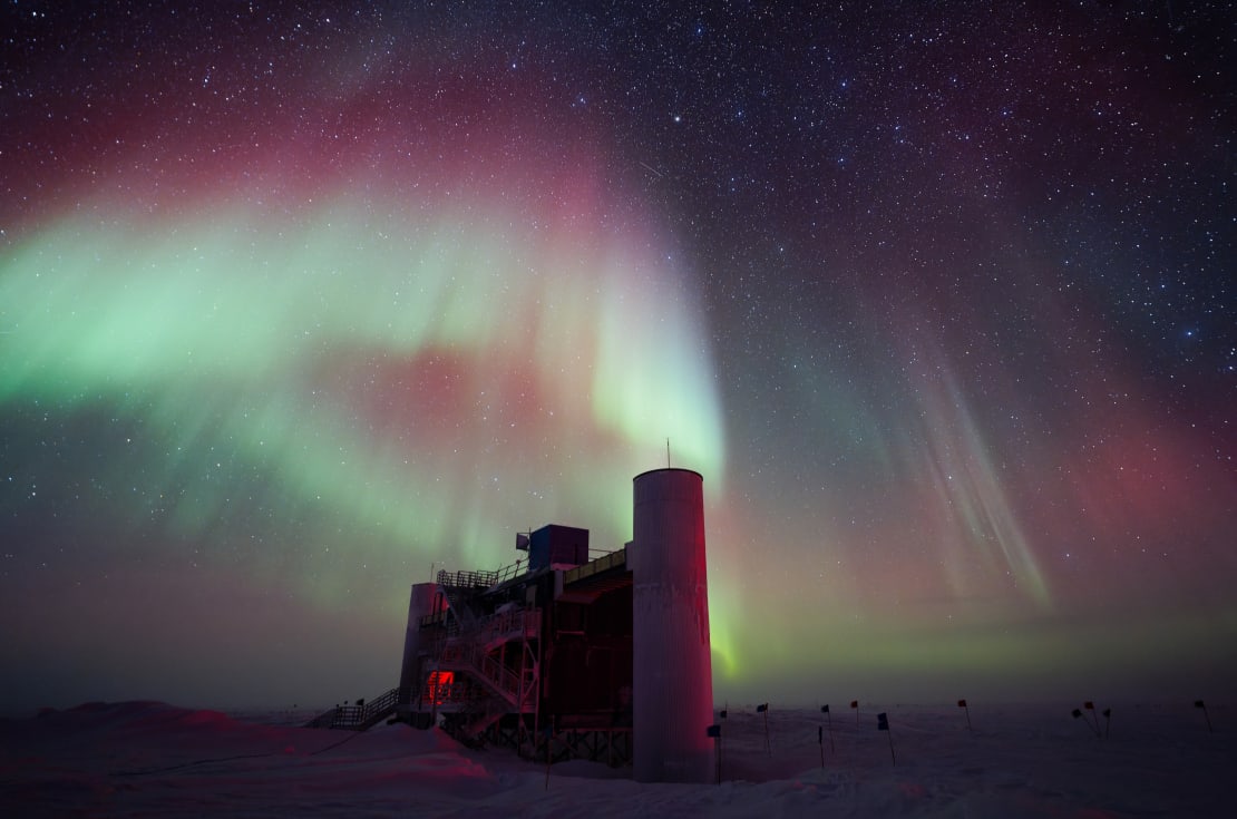 Multicolored auroras fanning out across sky above IceCube Lab, shown almost from side, in full shadow.